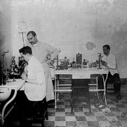 Imagem que mostra um ambiente de laboratório no prédio do Pavilhão Mourisco do antigo IOC. O auxiliar de laboratório, José Borges, sentado ao fundo, trabalha na preparação de lâminas. A imagem mostra ainda Carlos Chagas, sentado ao microscópio e Rocha Lima, de pé, ao seu lado. 