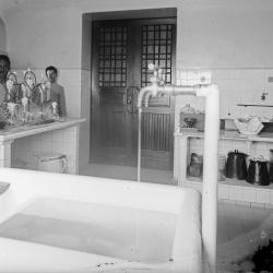 fotografia em preto e branco da sala de meios de cultura no Castelo. na frente da imagem, bastante desfocada, está a torneira de um aparato hidraulico, inventado por um dos cientistas do IOC, para produção de água destilada. ao fundo, uma bancada de azulejos com a vidraria e atrás da bancada, dois trabalhadores auxiliares em destaque. 