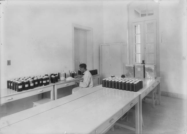 a imagem mostra uma sala de laboratório com uma grande bancada e um homem sentado que manipula drágeas de medicamentos, armazenando-as em um pote de vidro escuro. Sob a bancada há vários potes iguais aos que o homem está enchendo