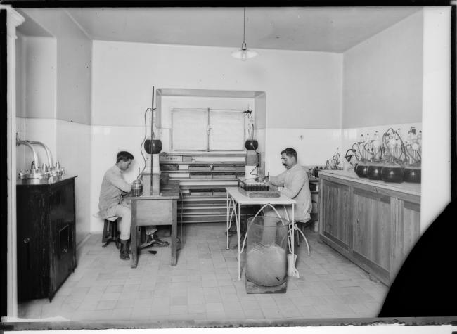 dois homens estão sentados em mesas posicionadas frente à frente em um laboratório. Em cima de cada mesa há um recipiente grande e esférico com um líquido escuro. Através de uma mangueira os auxiliares transferem o líquido para recipientes menores, também de vidro