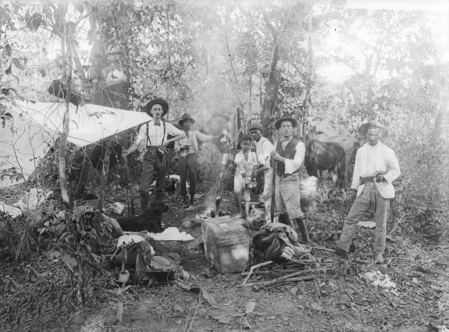 cena de um acampamento em uma mata ao centro, duas crianças e um homem preparam a comida