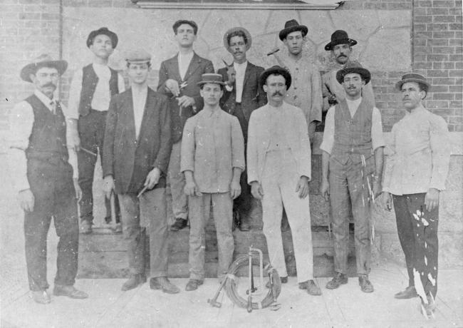 Fotografia em preto e branco de um grupo de onze trabalhadores especializados que trabalharam nas obras do Castelo Mourisco. Em frente a eles, no chão, estão suas ferramentas de trabalho.