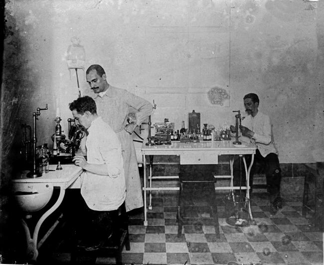 ambiente de laboratório. à frente, dois homens examinam algo no microscópio. ao fundo, um outro homem trabalha em uma mesa de bancada com um maçarico
