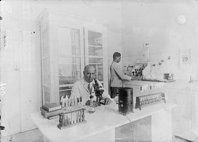 ambiente de laboratório. Na frente, um homem calvo trabalha em uma mesa em um microscópio. ao fundo, um outro homem está de pé em frente a uma pia com as vidrarias organizadas sob a bancada 