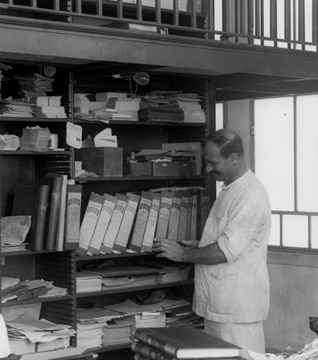 Um homem alto de bigodes junto a uma estante cheia de livros, dentro de uma biblioteca