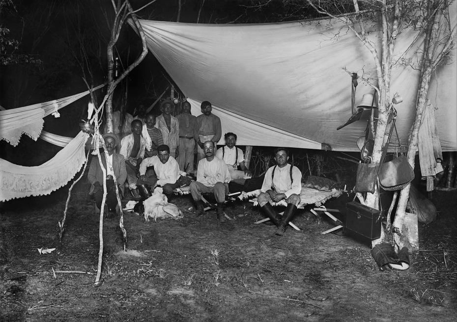 Acampamento da expedição de Belisário Penna e Artur Neiva. A imagem mostra um acampamento limpo e bem organizado. do lado direito, os cientistas arthur neiva e belisário penna estão sentados em cima de duas camas portáteis. do lado esquerdo está o grupo de auxiliares e algumas redes penduradas nas árvores.