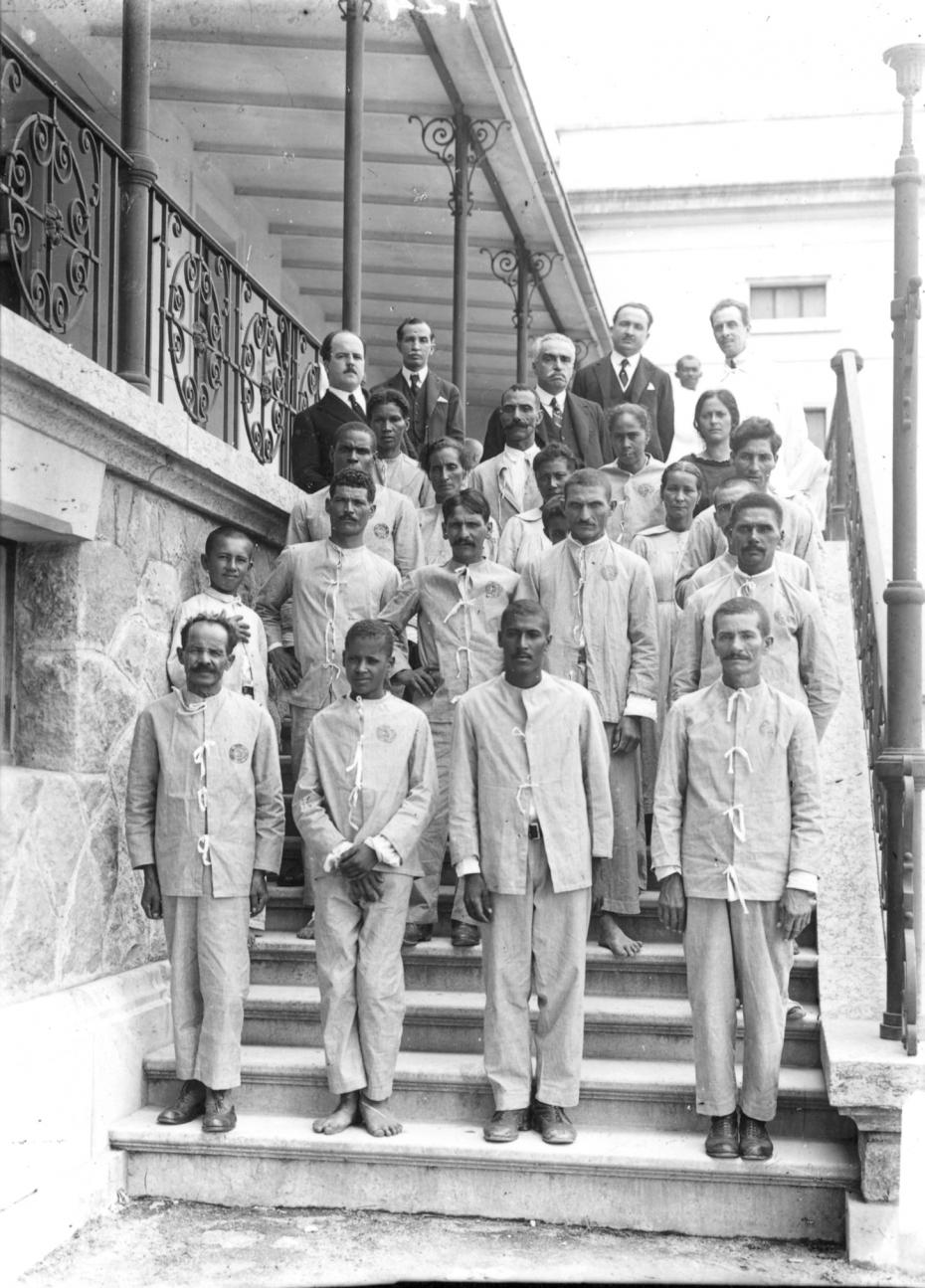 Foto tirada na escadaria do antigo prédio do INI. Nos degraus está um grupo de pacientes vestidos com roupa de internos do hospital. No alto, um grupo de médicos da academia nacional de medicina com Carlos Chagas. Altino está na parte mais alta da escada, segurando no corrimão