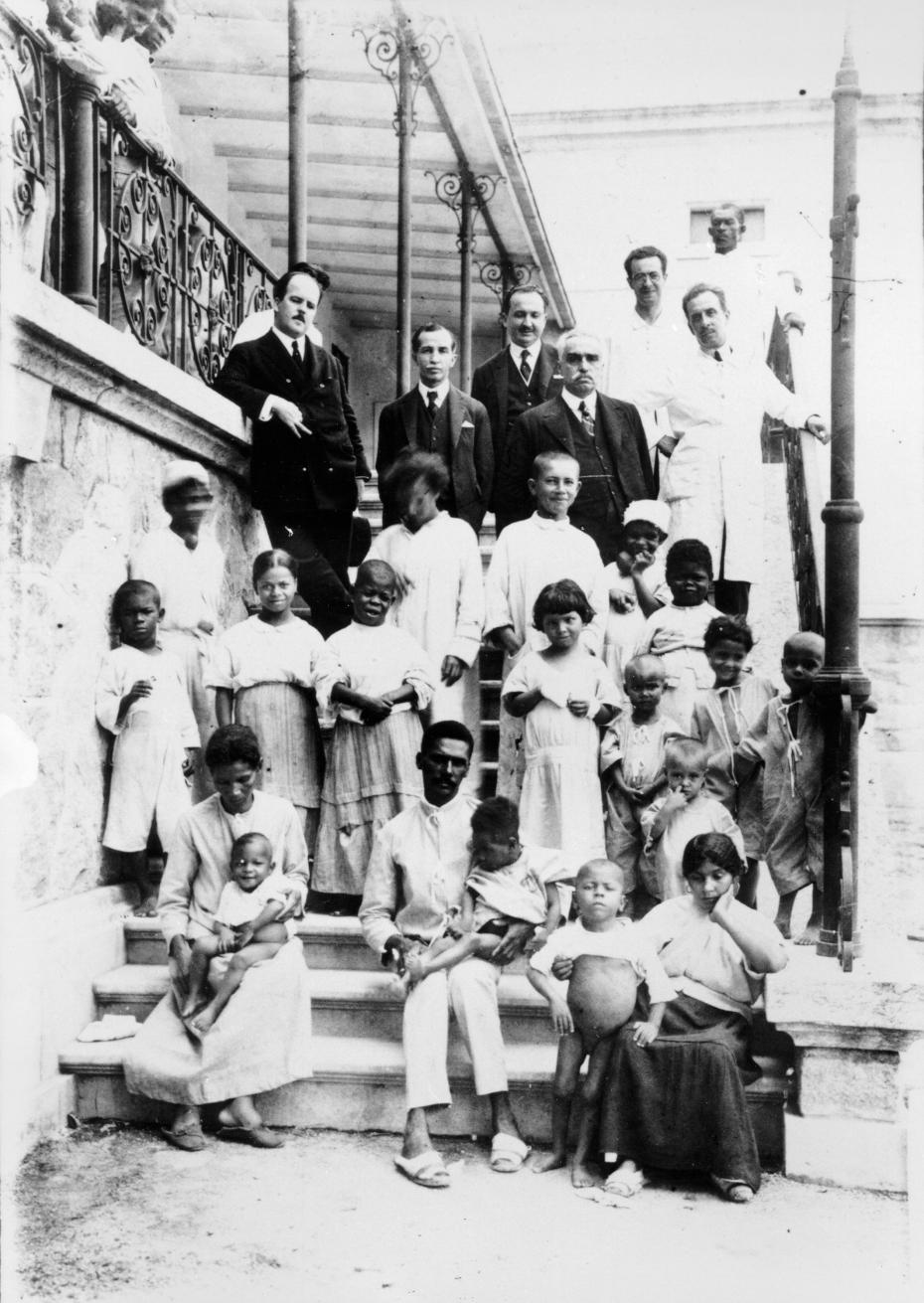 Foto tirada na escadaria do antigo prédio do INI. Nos degraus está um grupo de pacientes vestidos com roupa de internos do hospital. No alto, um grupo de médicos da academia nacional de medicina com Carlos Chagas. Altino está na parte mais alta da escada, segurando no corrimão