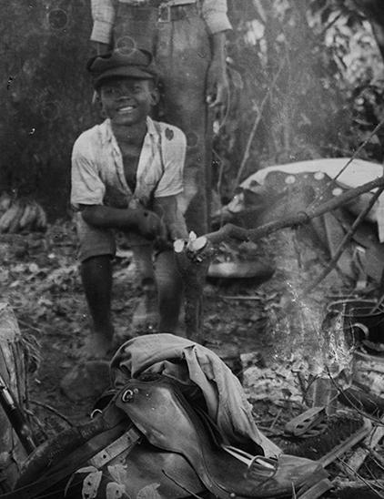 um menino sentado em um acampamento na mata com um fogareiro e panelas ao seu lado