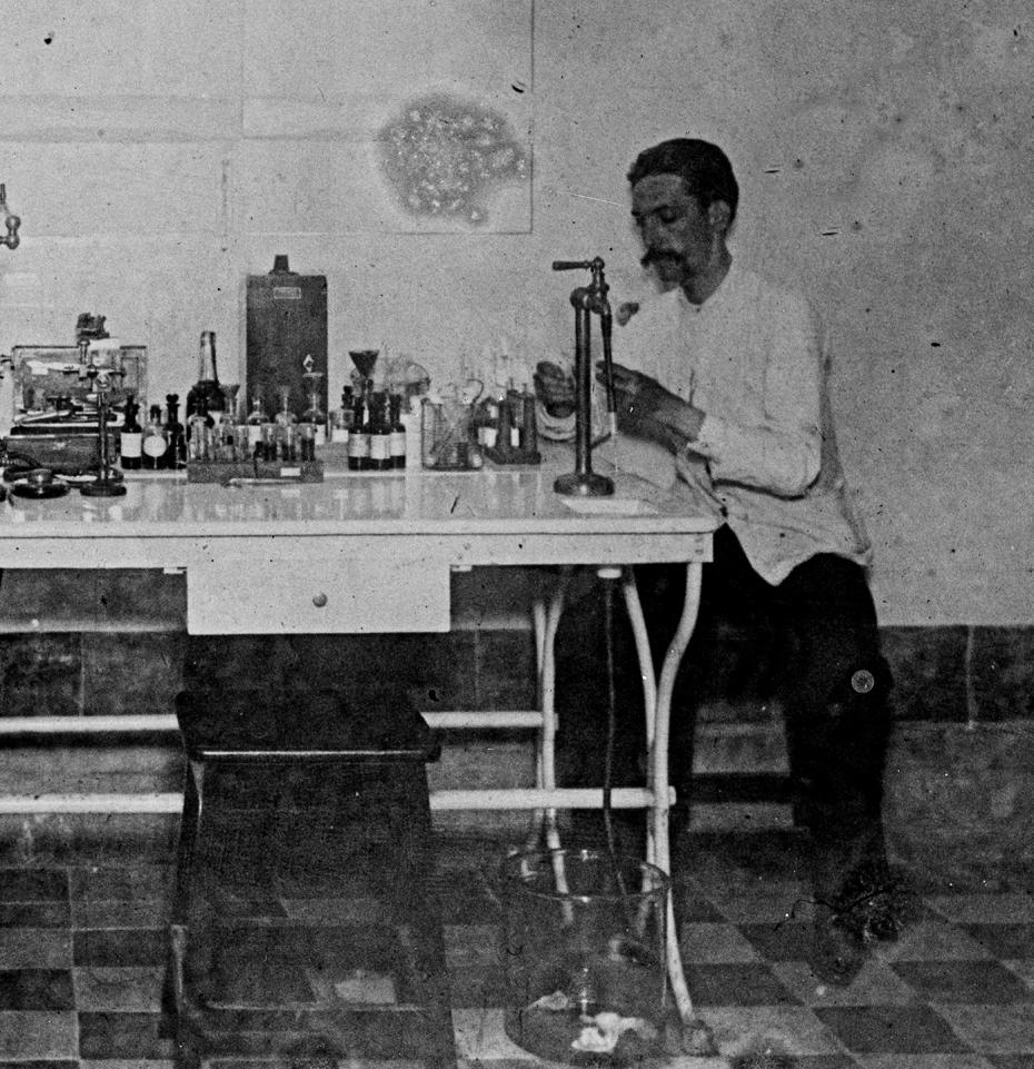 José Borges auxiliar de laboratório, trabalha em uma mesa no laboratório de Carlos Chagas. Vestido de jaleco, usa um longo bigode à moda da época