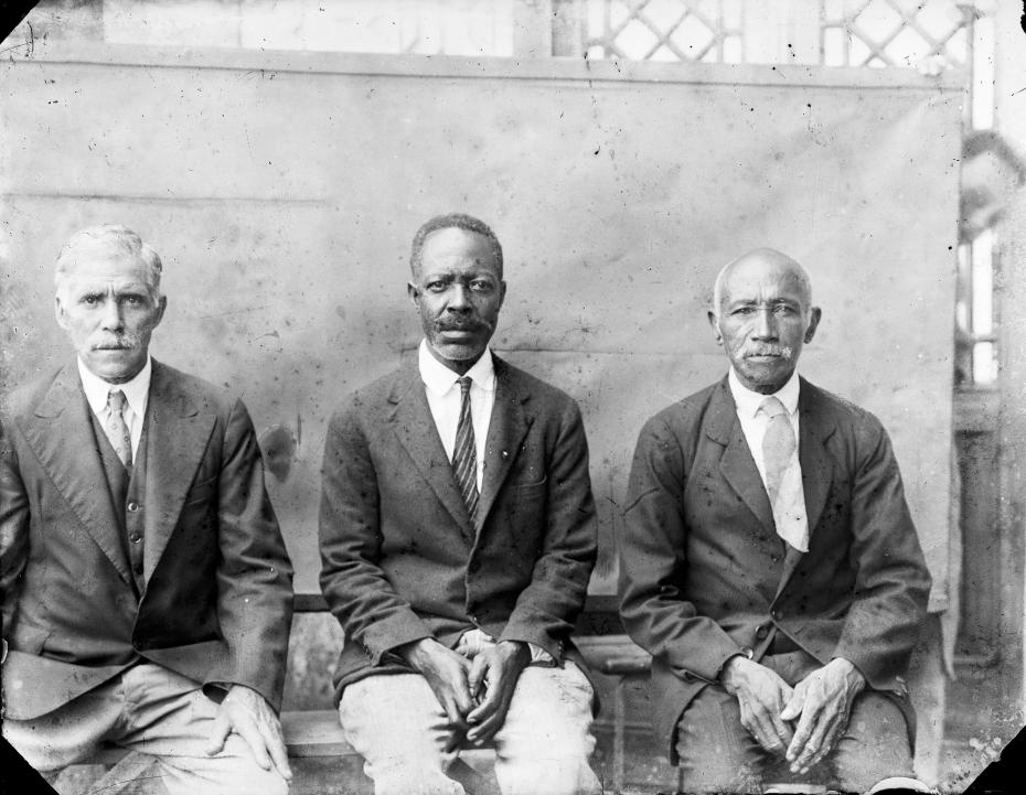 três homens sentados posando para uma foto. os três estão vestidos de paletó e gravata