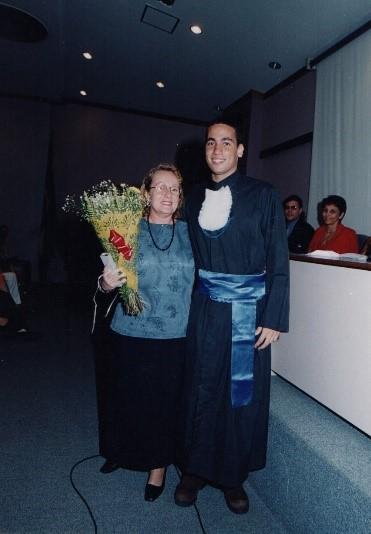 Um aluno vestido de beca abraçado com sua professora que segura um buquê de flores